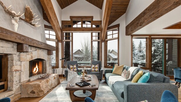 Amplia sala de estar con chimenea de piedra, vigas de madera y ventanas grandes que ofrecen vista a árboles cubiertos de nieve.