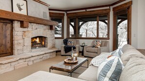 Un salón acogedor con chimenea de piedra, asientos cómodos y ventanas amplias que ofrecen vistas a árboles cubiertos de nieve.