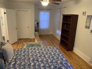 A bedroom with a bed, a wooden floor, a ceiling fan, a window with curtains, a bookshelf, and a door.