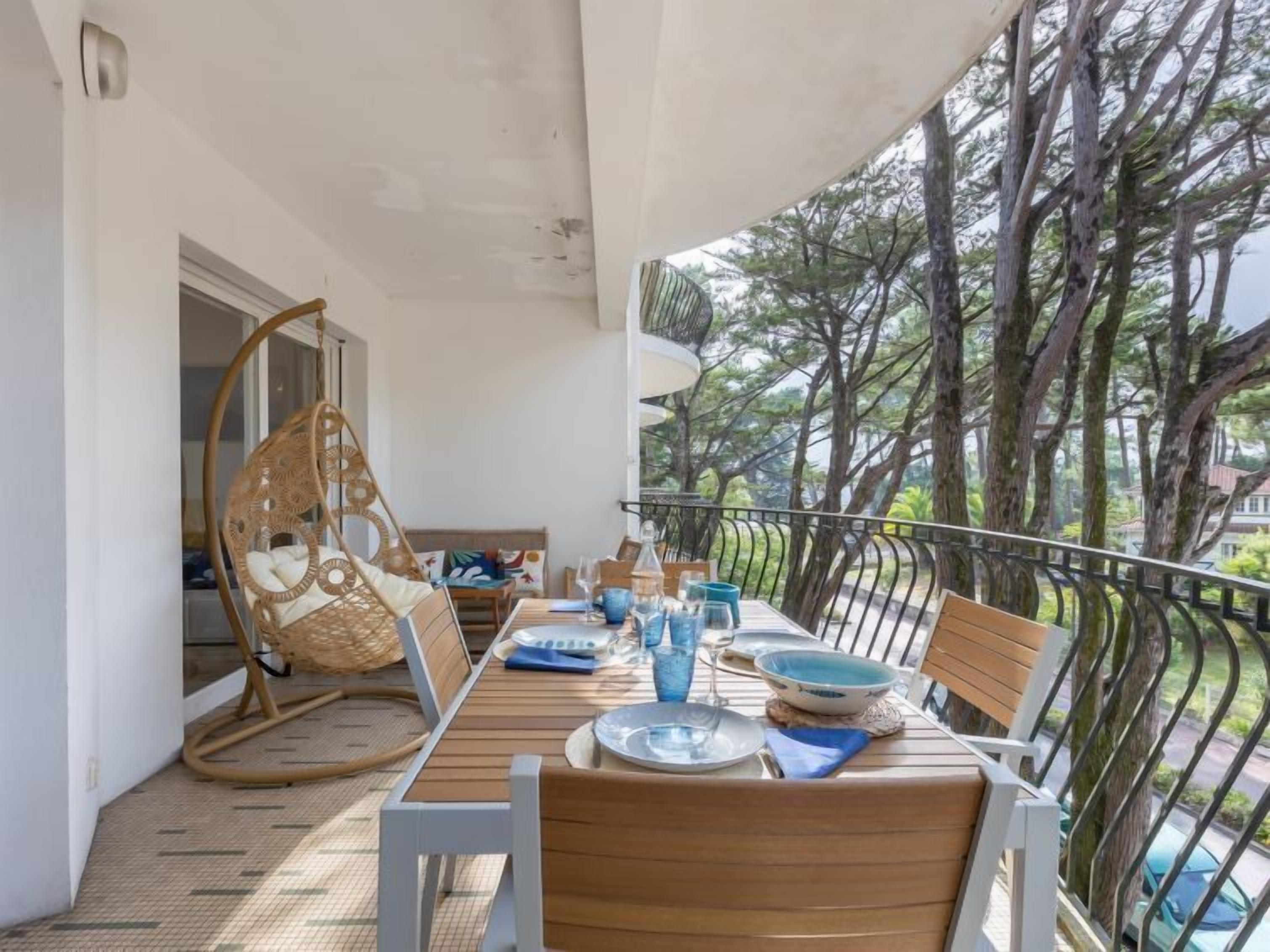 A balcony with a wooden table set for dining, a wicker chair, and a view of trees and buildings.