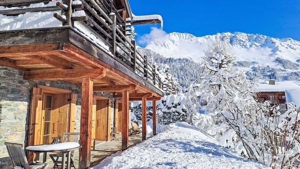 Una cabaña de madera con zócalo de piedra, porche de madera y vista a montañas nevadas.