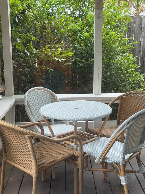 Un espacio de estar al aire libre con muebles de mimbre, una mesa redonda y sillas, rodeado por una terraza de madera y una barandilla blanca.