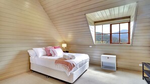 Een slaapkamer met een schuine kap, een bed met witte beddengoed en roze kussens, een nachtkastje met een lamp en een raam met uitzicht op bergen.
