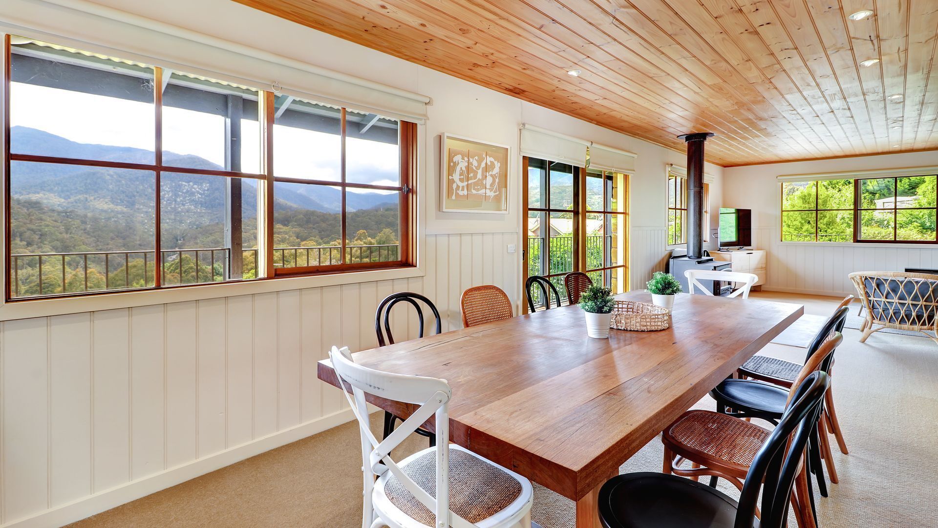 A spacious dining area with a long wooden table, white and wicker chairs, and large windows offering a mountain view.