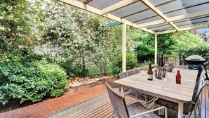 Een terras buiten met een houten tafel en stoelen, omgeven door weelderige begroeiing en een metselpad.