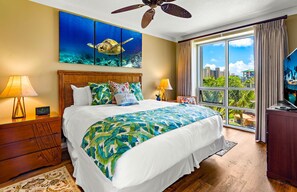 Une chambre d’hôtel avec un lit, un ventilateur de plafond, une grande fenêtre donnant sur la ville et un tableau représentant une tortue de mer.