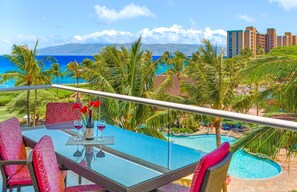 Un balcon avec une table dressée pour deux personnes, donnant sur une piscine et offrant une vue sur l’océan et les bâtiments.