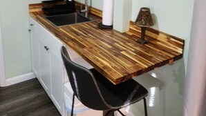 A compact kitchen with a wooden countertop, a sink, and a small dining area with a chair. There are shelves with decorative items and a wall-mounted sign. - Centrally Located Happy Haven (San Antonio)