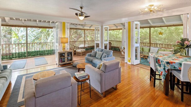 A spacious living area with a wooden floor, a sofa, a coffee table, and a dining table with a floral tablecloth. - Molokai Oceanfront large beautiful home - Gorgeous secluded beaches out front! (Maunaloa)