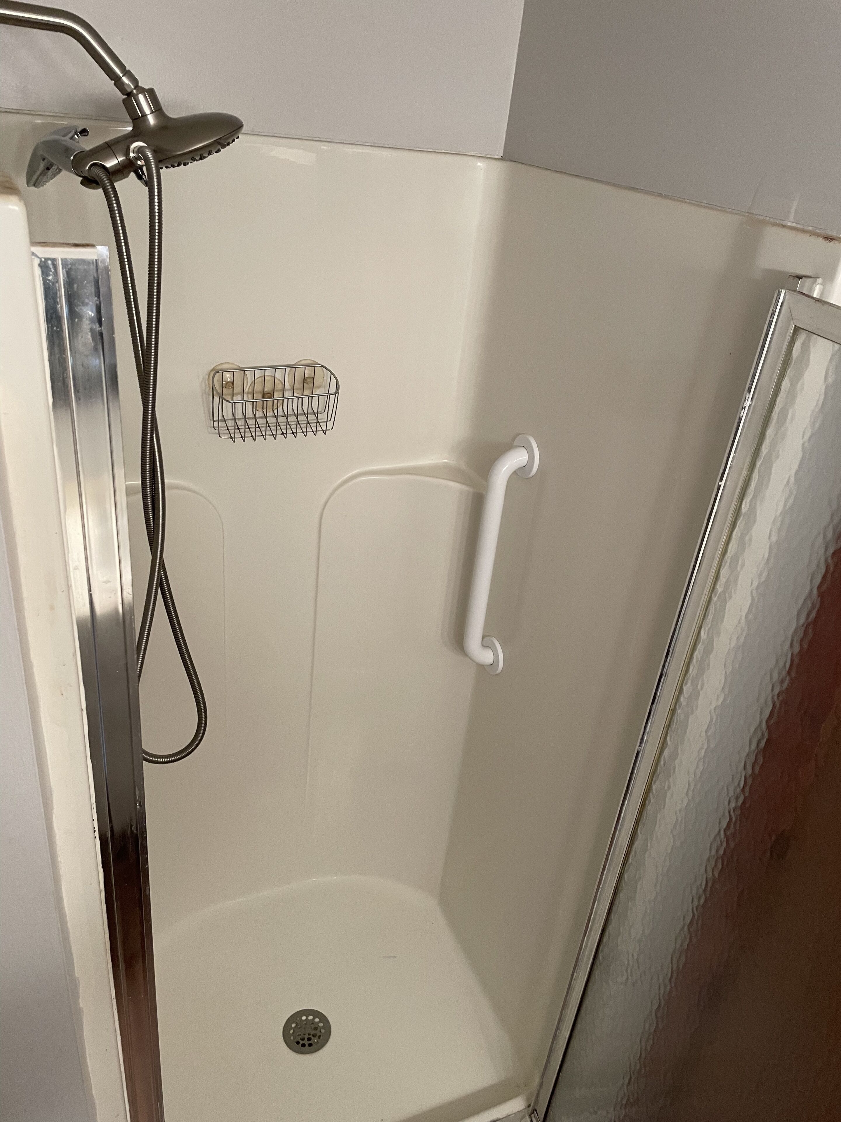 Une douche équipée d’un pommeau de douche à main, d’une barre d’appui blanche et d’une étagère de douche en métal.