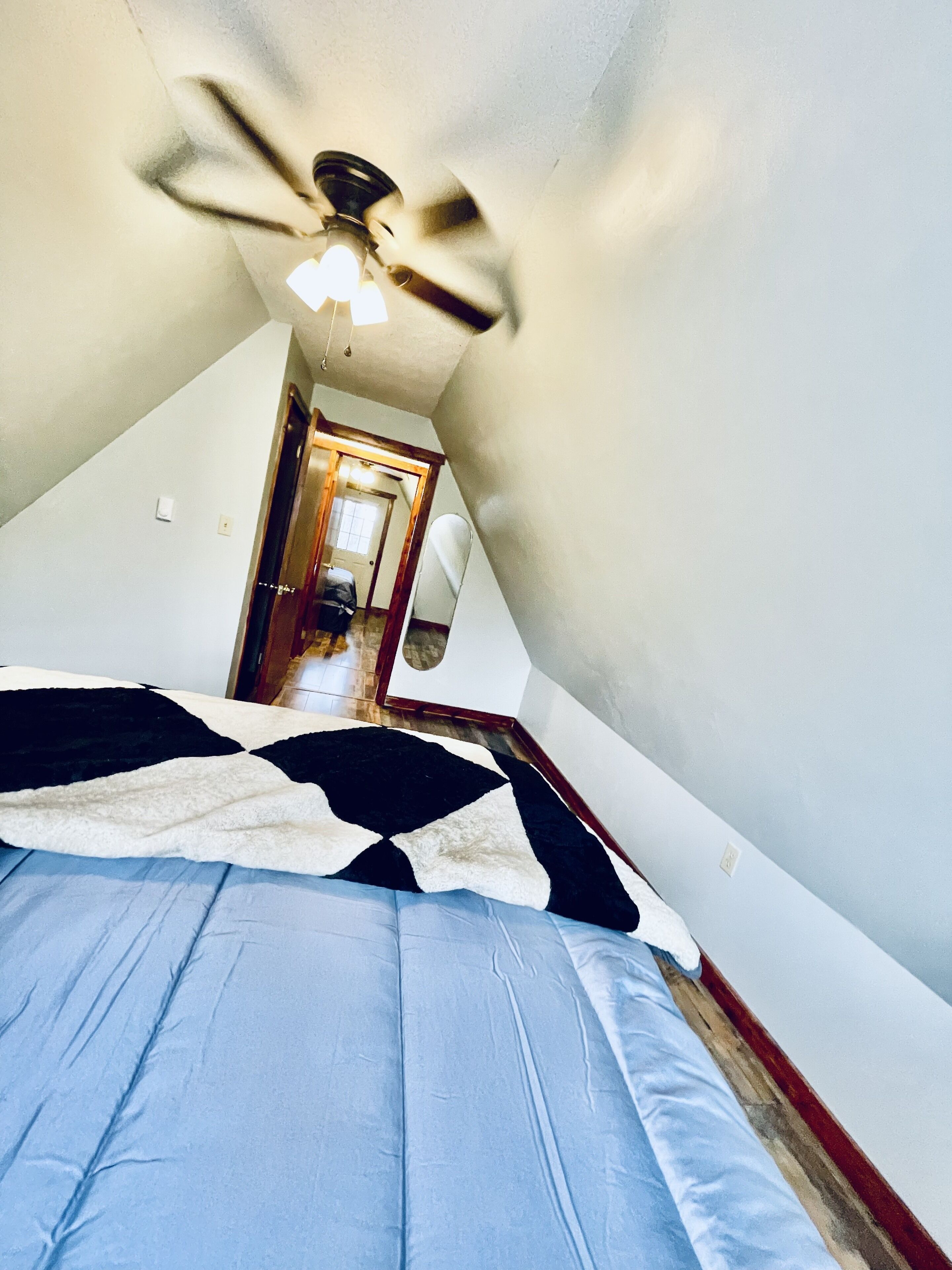 Une chambre avec un plafond en pente, un ventilateur de plafond, un lit recouvert d’une couverture à motifs et une porte visible donnant sur une autre pièce.