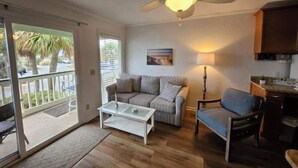 Una sala de estar con ventilador de techo, un sofá, una mesa de centro, una silla y una cocina con armarios.