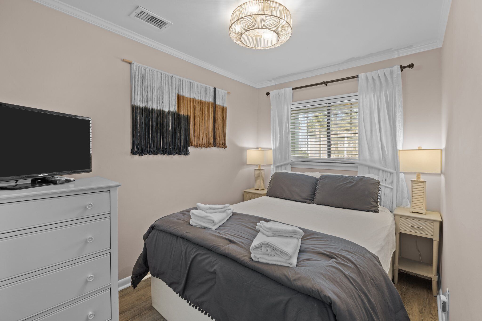 Un dormitorio ordenado con cama, mesitas de noche, televisor y ventana con cortinas.