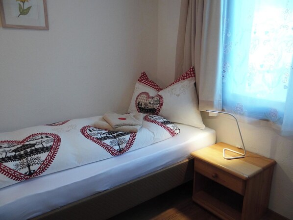 Una cama individual con colchón blanco y ropa de cama con estampado de corazones, una mesita de noche con una lámpara y una ventana con cortina.