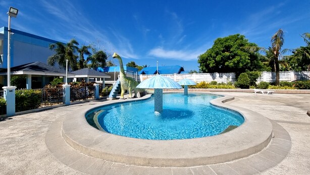 Une piscine agrémentée d’une statue de dinosaure et de parasols, entourée d’une zone pavée et de palmiers.