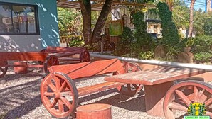 Un espace extérieur aménagé dans un style rustique, avec des bancs en bois et des roues, entouré de verdure et agrémenté d’une pergola.