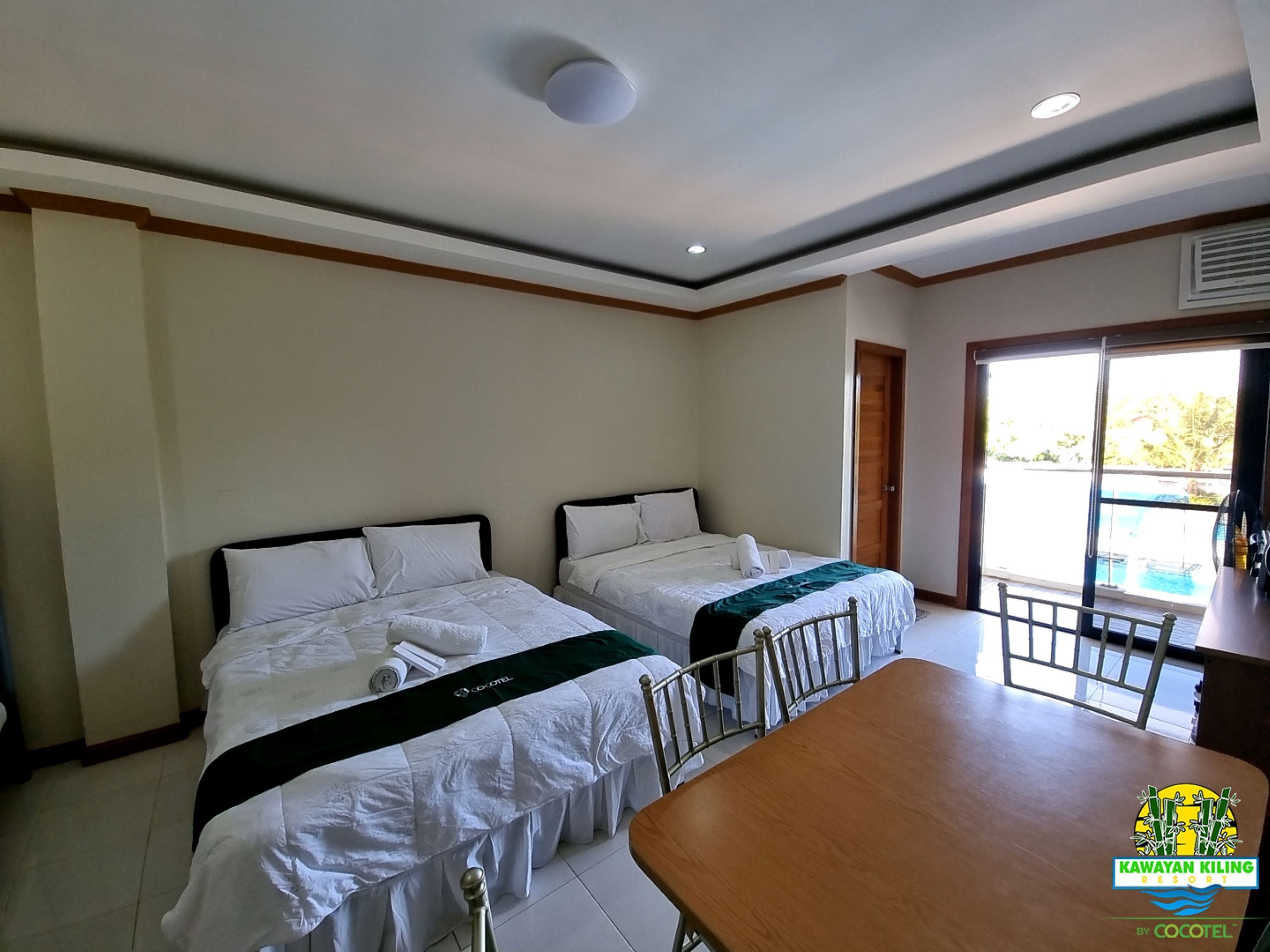 Habitación de hotel con dos camas, una mesa de comedor de madera y una puerta corrediza de vidrio que da a una zona de piscina.