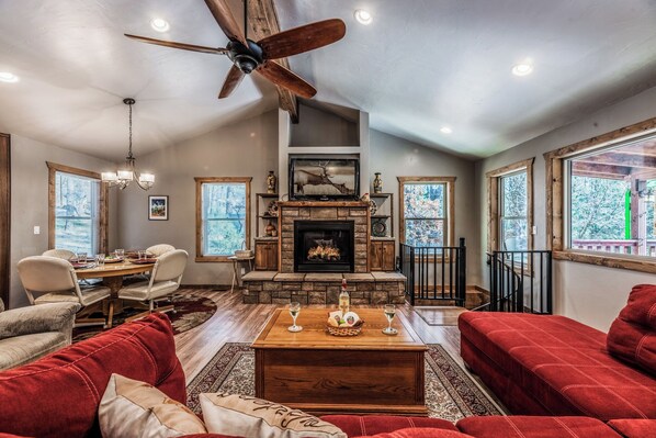 A spacious living room with a fireplace, red sofas, a dining area with a table and chairs, and large windows with a view of trees. - The Great Escape by Ruidoso Vacation Rentals (Ruidoso)