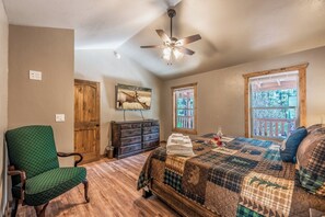 A bedroom with a bed, a green chair, a wooden dresser, a ceiling fan, and a window with a view of a balcony. - The Great Escape by Ruidoso Vacation Rentals (Ruidoso)