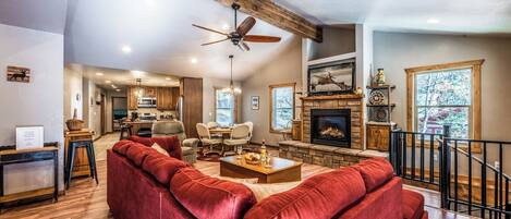 A spacious living room with a red sofa, wooden ceiling beams, a fireplace, and a kitchen in the background.