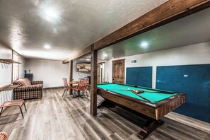 A pool table in a room with wooden flooring and furniture, including a sofa and chairs. - The Great Escape by Ruidoso Vacation Rentals (Ruidoso)