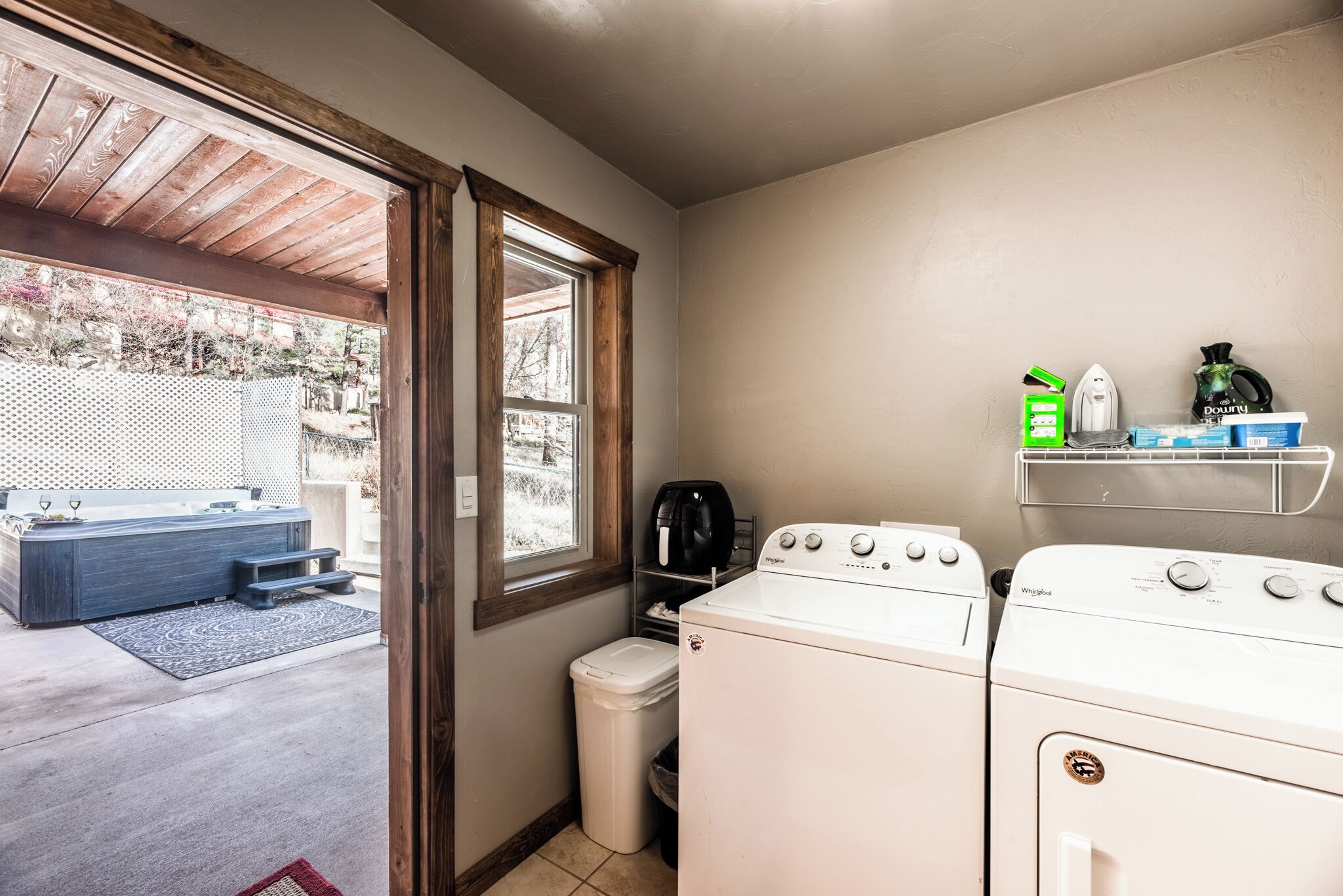 A laundry area with two white washers, a small trash bin, and a window with a view of a backyard.