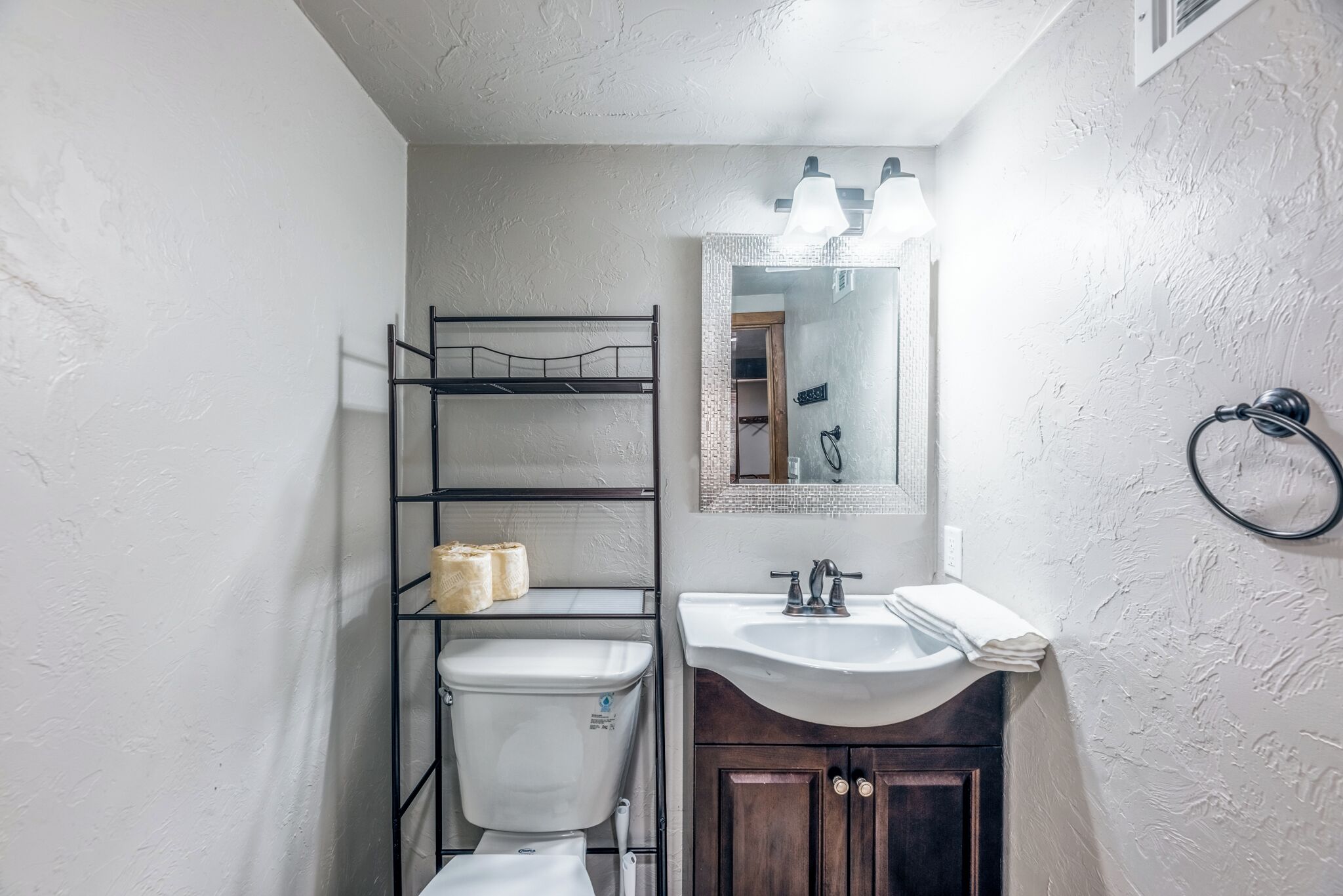 Ein kleines Badezimmer mit Toilette, Waschbecken und einem Spiegel darüber. Eine Ablagefläche mit Toilettenartikeln und ein Handtuchhalter sind an der Wand angebracht.