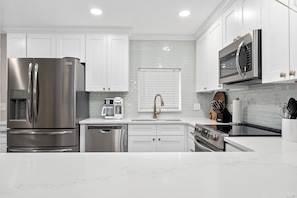 A modern kitchen with stainless steel appliances, white cabinetry, and a marble countertop. - Trade Winds 101 by Distinctive Beach Rentals (St. Augustine)