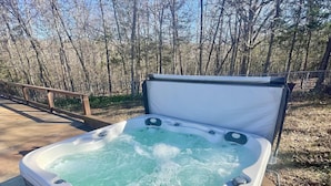 A hot tub with a control panel and a smartphone on it, surrounded by a wooden deck and a metal railing, with trees in the background.
