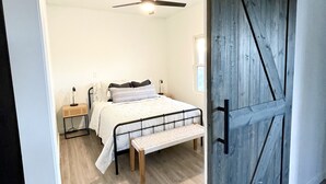 A bedroom with a bed, a bench, a bedside table, a ceiling fan, and a wooden door.
