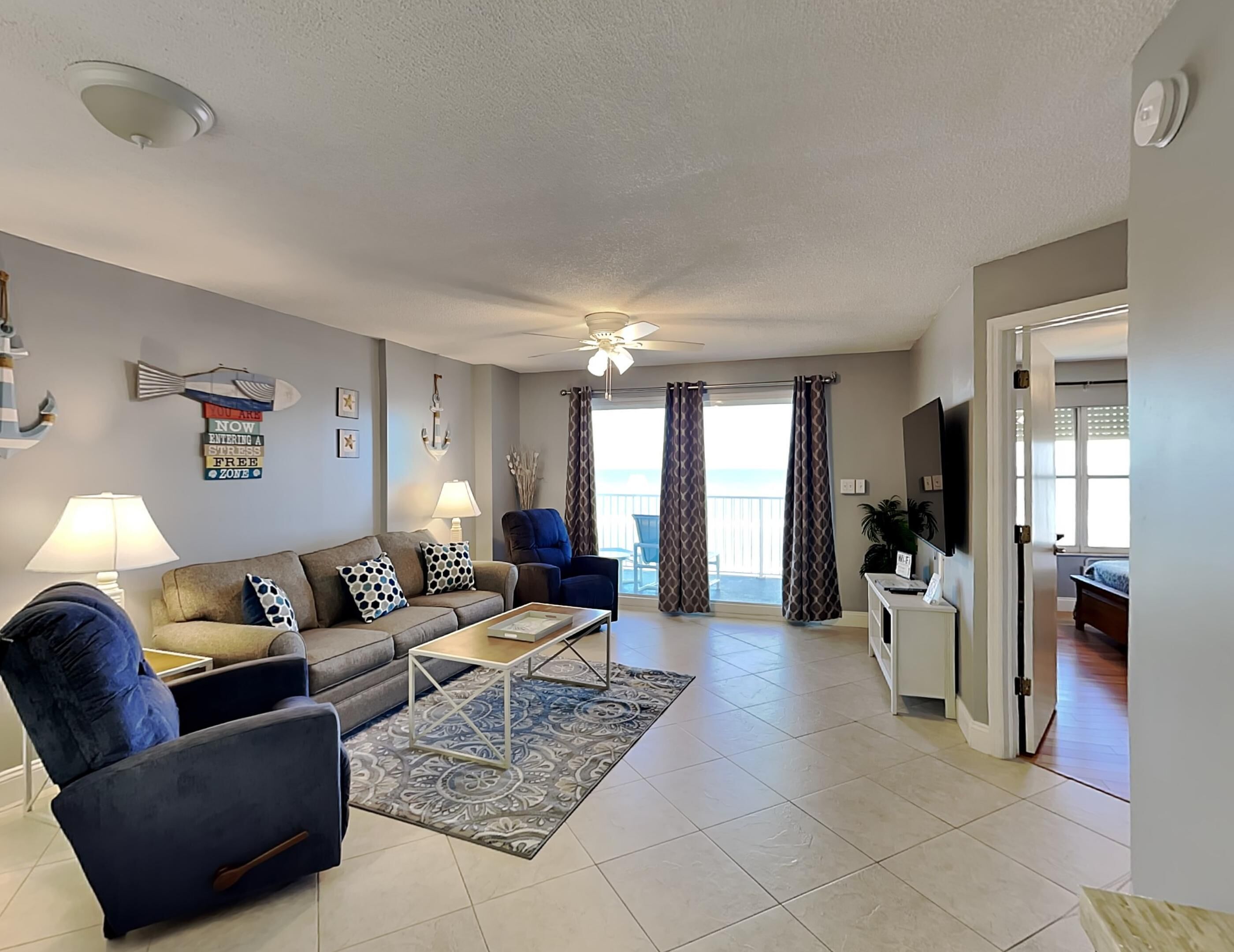 A living room with a sofa, armchairs, a coffee table, and a TV. There is a balcony with curtains and a view of the outdoors.