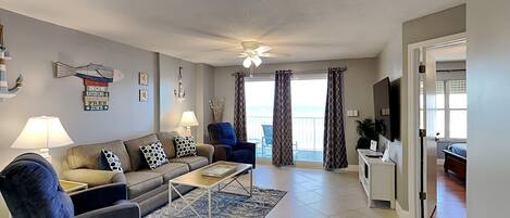A living room with a sofa, armchairs, a coffee table, and a TV. There is a balcony with curtains and a view of the outdoors.