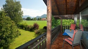 Un balcone in legno con tavolo e sedie, affacciato su un paesaggio erboso con alberi e montagne sullo sfondo.