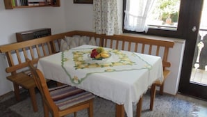 Een eethoek met een tafel bedekt met een witte tafelkleed met bloemenprint, een fruitschaal en een houten bank.