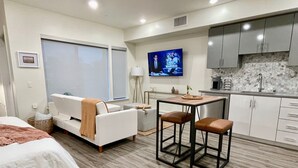 A modern hotel room with a bed, a sofa, a small dining table with two stools, a TV, and a kitchen area with cabinets and a sink. - Pets Stay Free in Downtown Walnut Creek Condo (Walnut Creek)