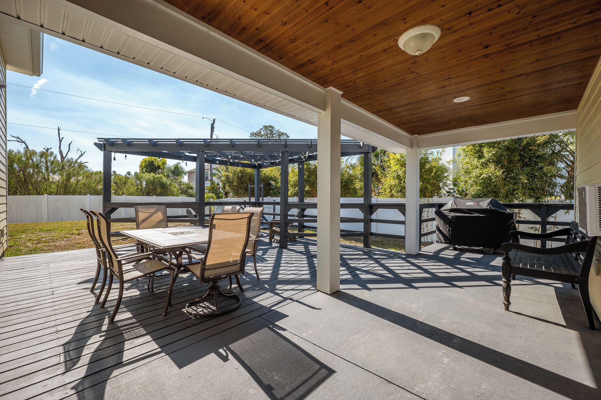 En overdækket udendørs terrasse med træloft, pergola og et bord med stole samt grill.