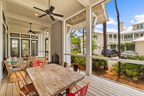 Um deck coberto ao ar livre com mesa e cadeiras de madeira, janelas amplas e ventilador de teto.