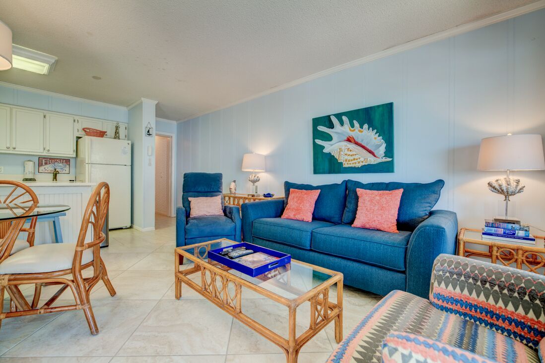 A living room with a blue sofa, a coffee table, and wicker chairs. There is a kitchen area with white cabinets and a refrigerator.