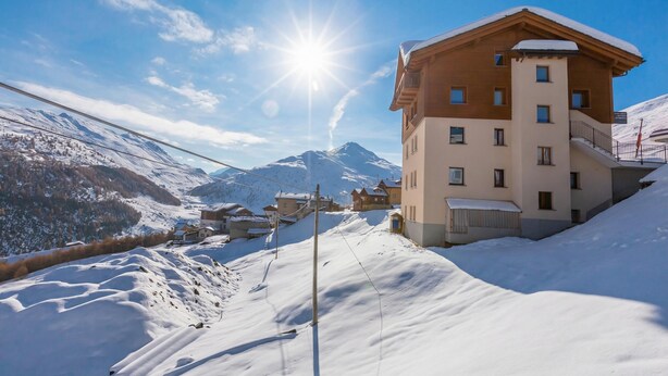 A snow-covered mountain village with a prominent building featuring multiple windows and a balcony. - Fiocco Di Neve Ski in- Ski out Mt.10 (Livigno)