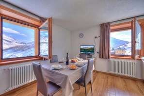 A dining area with a table set for two, a clock on the wall, and a view of a snowy landscape through large windows. - Fiocco Di Neve Ski in- Ski out Mt.10 (Livigno)