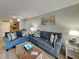 A living room with a sofa, coffee table, and a lamp. There is a kitchen in the background.