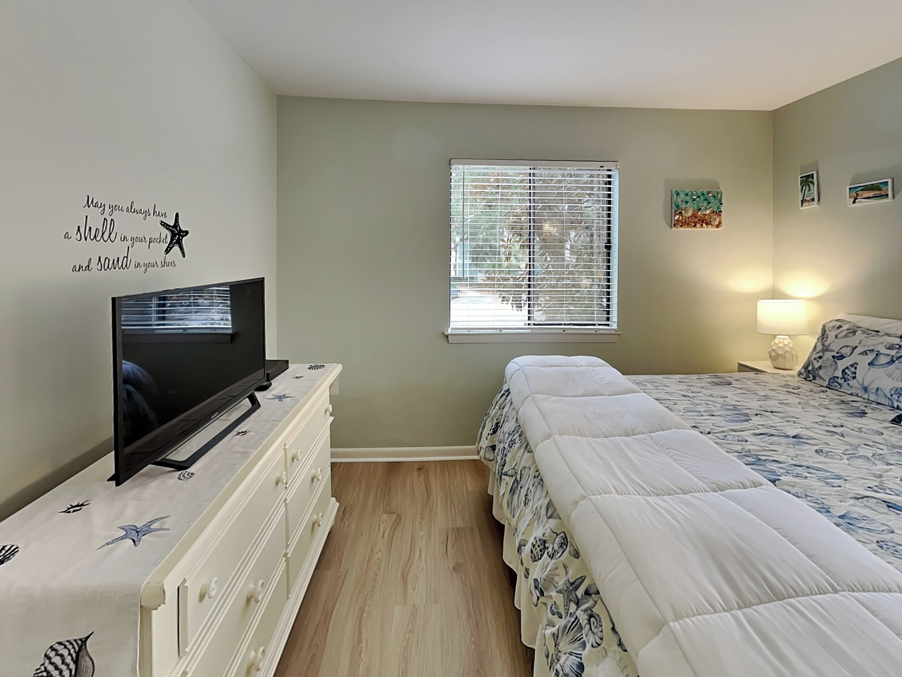 Un dormitorio con una televisión sobre un tocador, una ventana con persianas, una cama con edredón estampado y una pared con una cita decorativa.