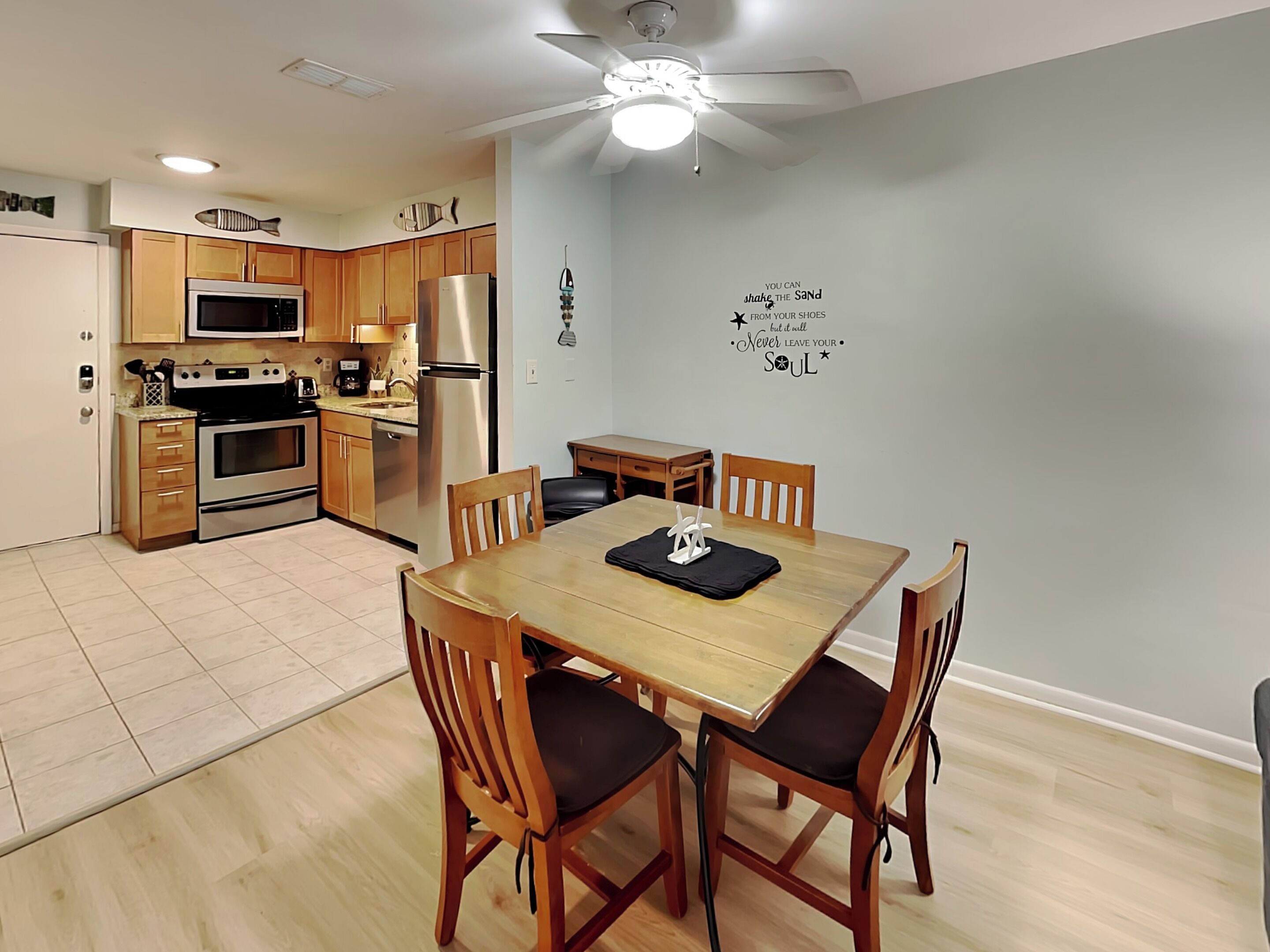 A modern kitchen with wooden cabinets, a stainless steel refrigerator, and a microwave. There is a dining table with four chairs and a ceiling fan.