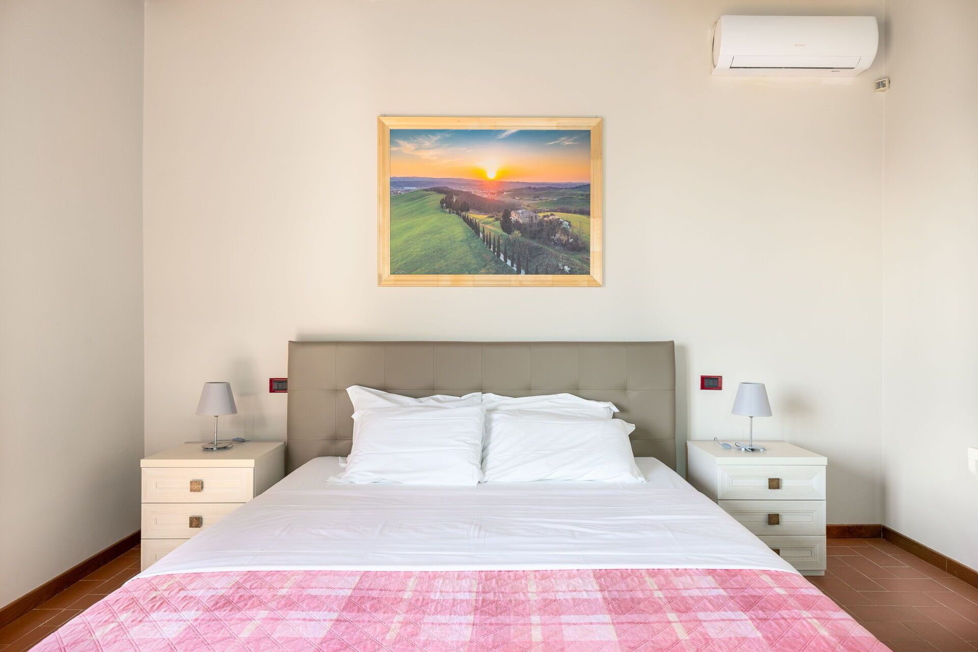 Une chambre d’hôtel comprenant un lit, deux tables de chevet, une commode, un climatiseur fixé au mur et un tableau représentant un paysage, encadré.