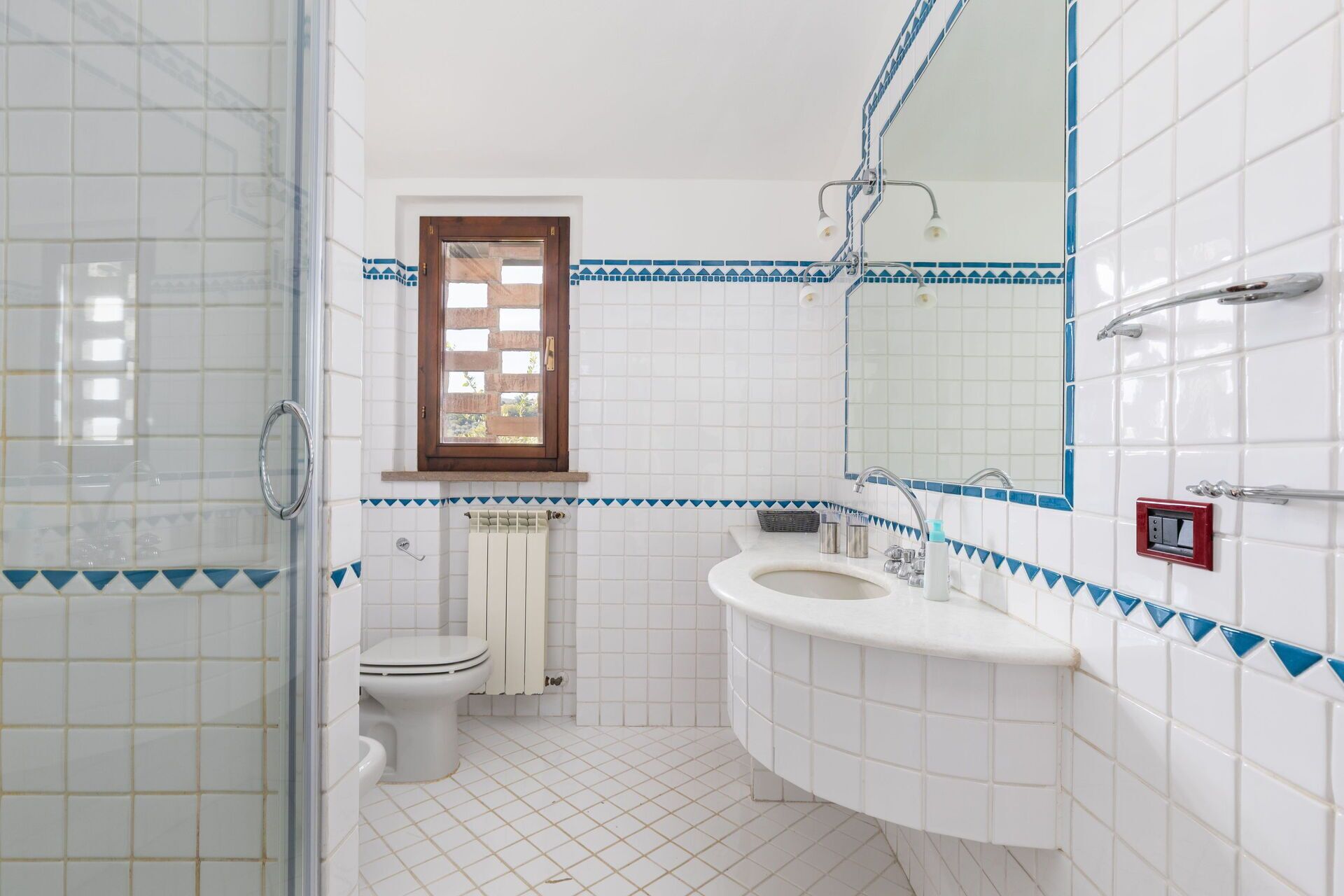 A bathroom with white tiles, a toilet, a sink, a shower, and a window with a wooden frame.