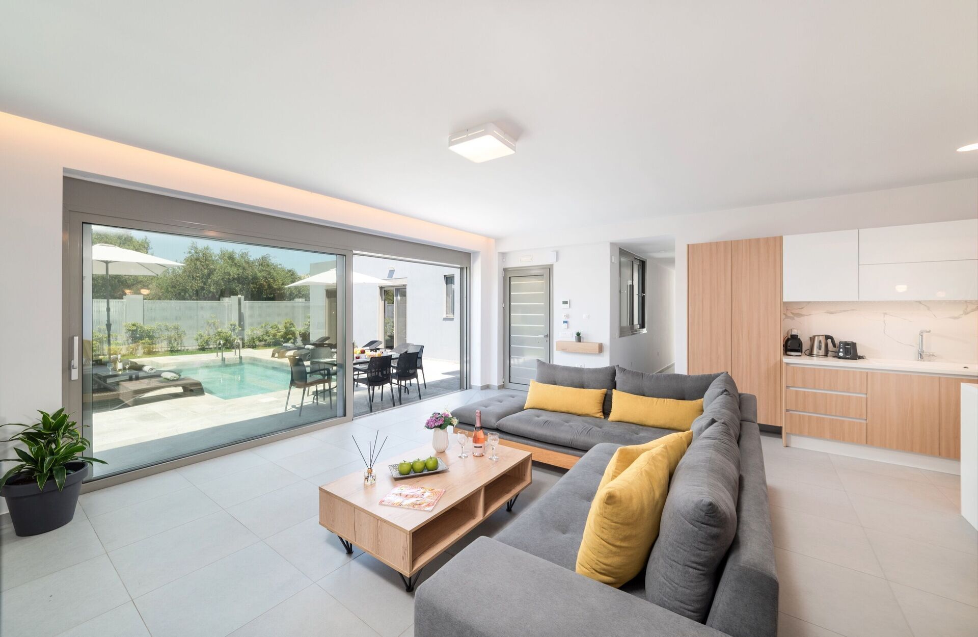 A modern living room with a grey sofa, a wooden coffee table, and a glass sliding door leading to a pool area.