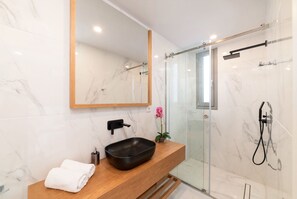 Une salle de bain moderne avec un meuble-lavabo en marbre, un lavabo noir, un plan de toilette en bois, une serviette blanche, un miroir, une cabine de douche en verre et une douchette à main.