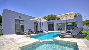 A modern pool area with lounge chairs and umbrellas, surrounded by a house with large windows and a pool deck.