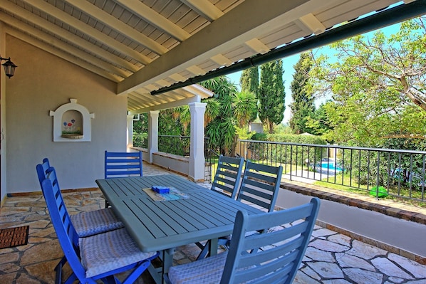 Un patio cubierto con una mesa y sillas, un adorno en la pared y vistas a una piscina y follaje.