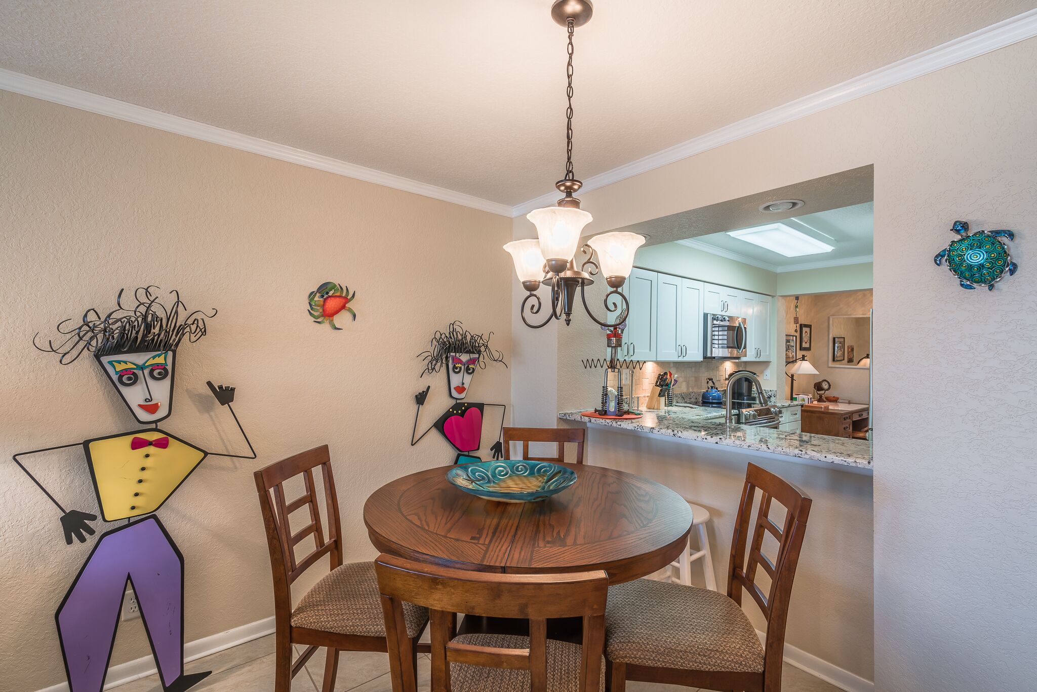 A dining area with a round wooden table, four chairs, a chandelier, and a kitchen visible through an open doorway.